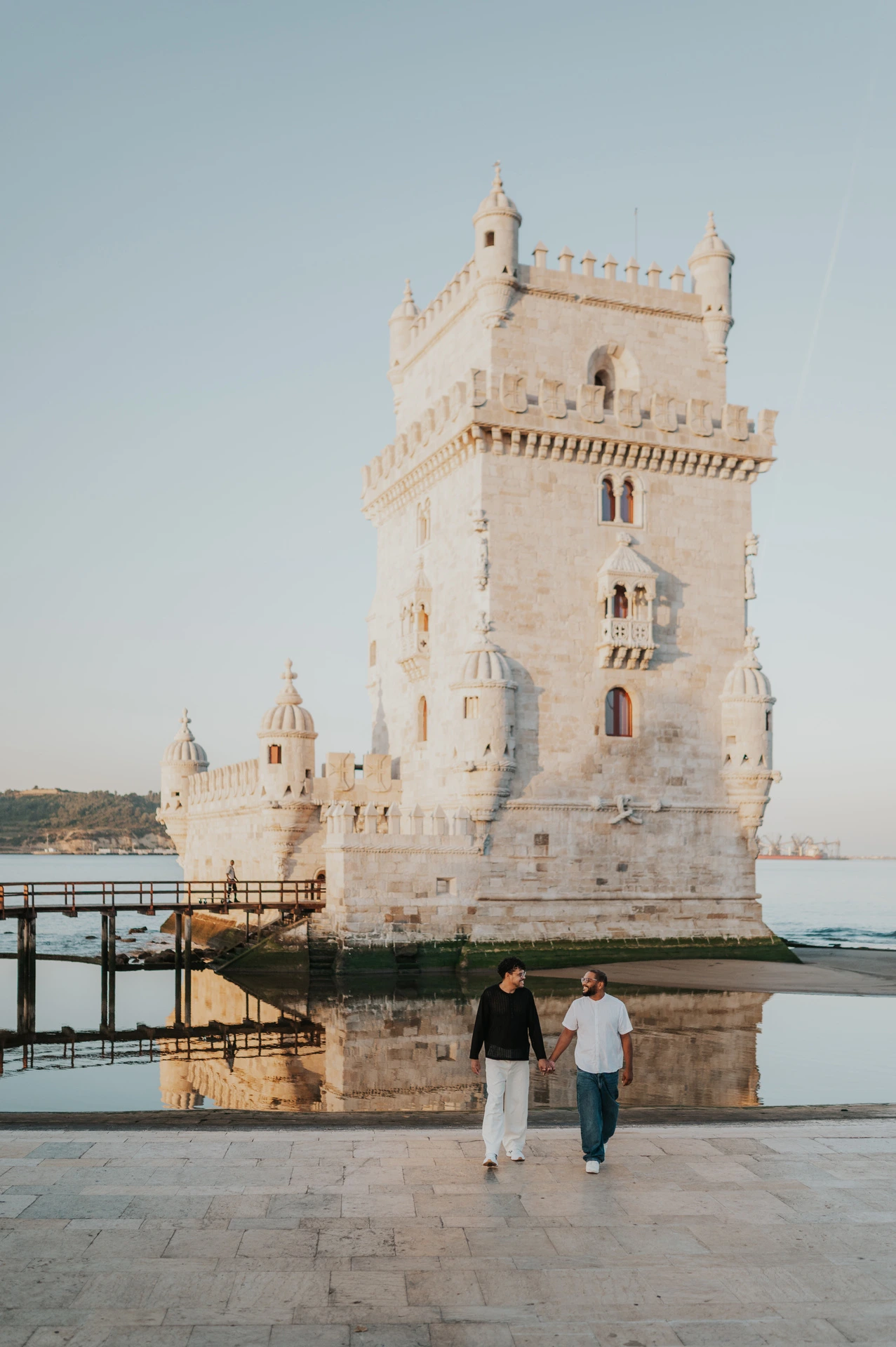 The couple walking towards the camera with Belém Tower and the water behind them during their couple photoshoot in Lisbon.