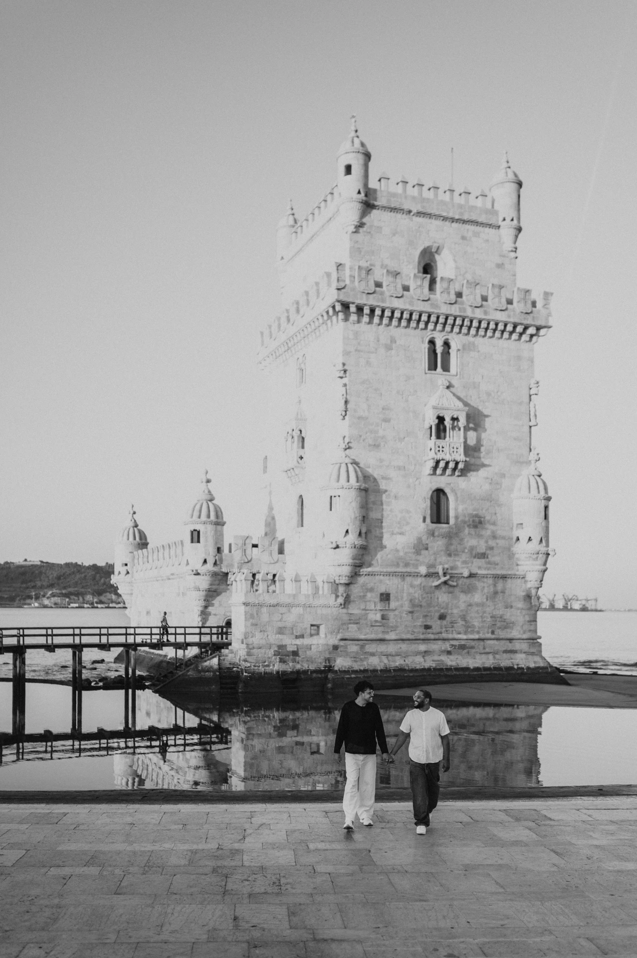 A black-and-white photograph of the couple walking hand in hand in front of Belém Tower during their couple photoshoot in Lisbon.