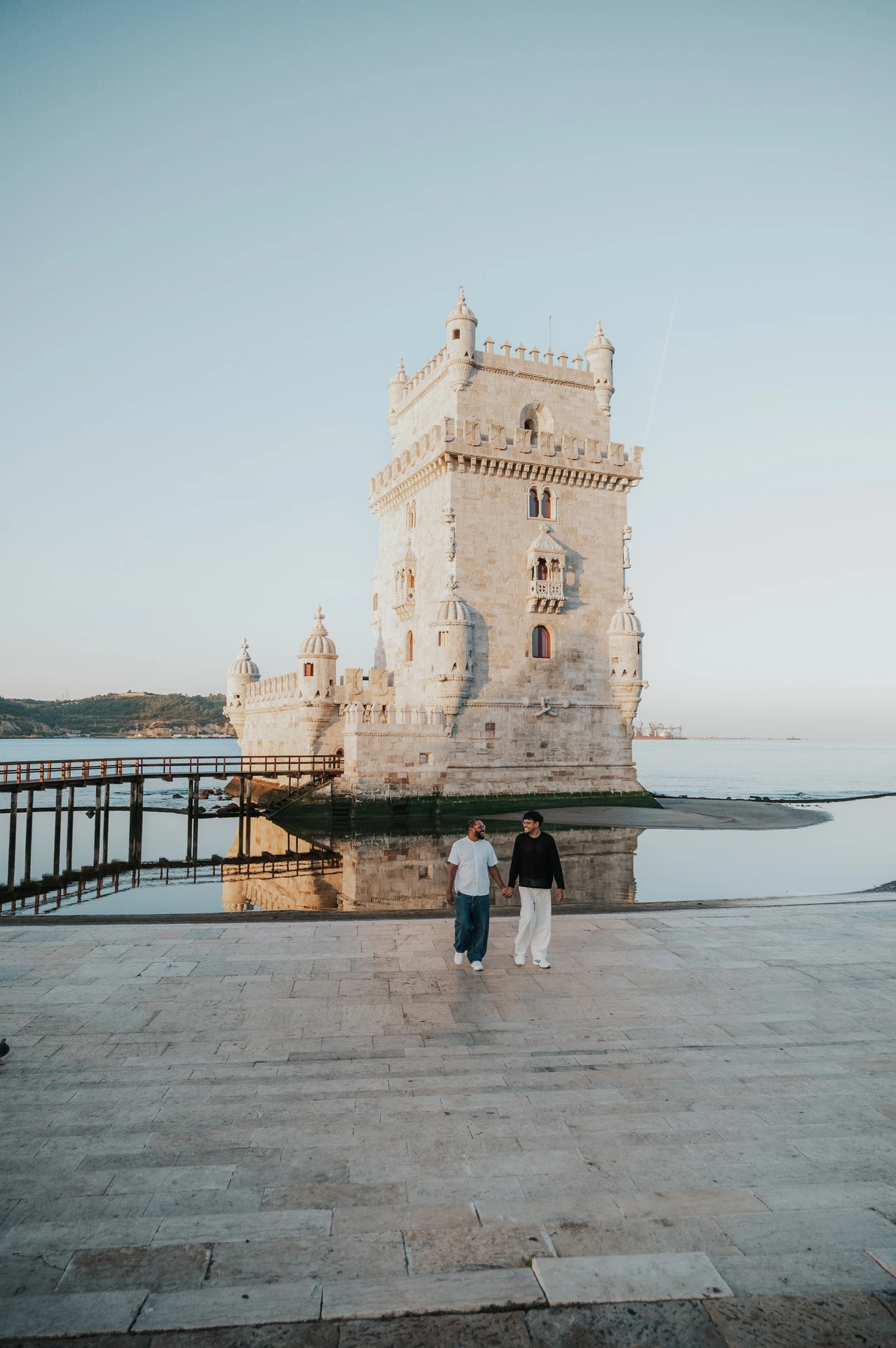 A distant view of the couple walking hand in hand across the stone paving in front of Belém Tower during their couple photoshoot in Lisbon.