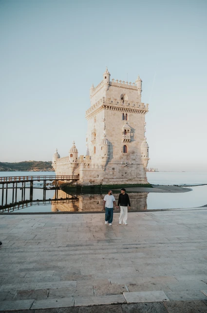 A wide view of their tower walk | Lisbon couple photoshoot A distant view of the couple walking hand in hand across the stone paving in front of Belém Tower during their couple photoshoot in Lisbon.