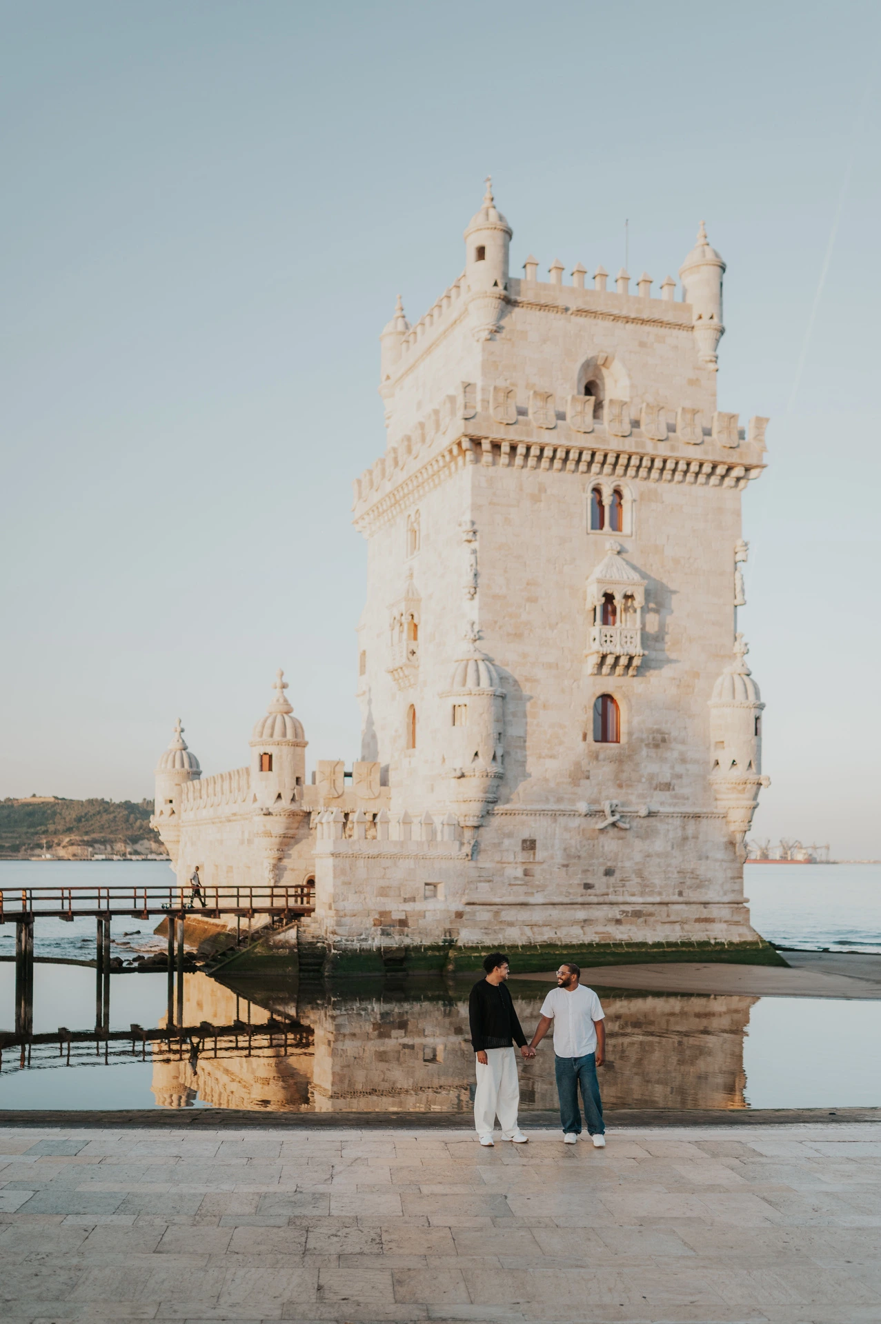 The couple holding hands as they walk beneath a full view of Belém Tower during their couple photoshoot in Lisbon.