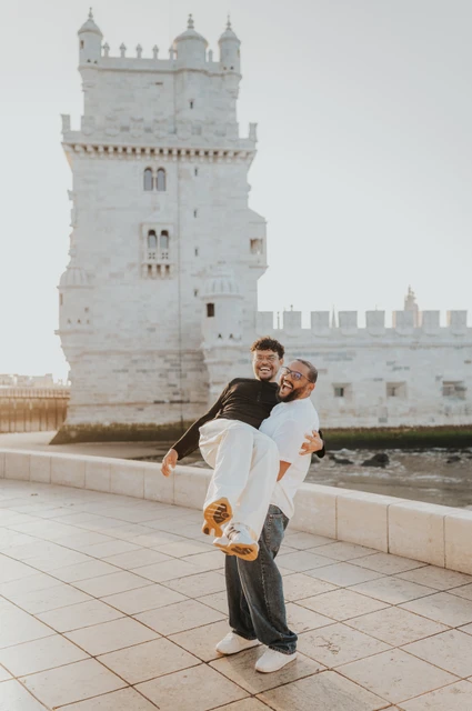 The couple smiling as one partner carries the other in his arms in front of Belém Tower during their couple photoshoot in Lisbon.