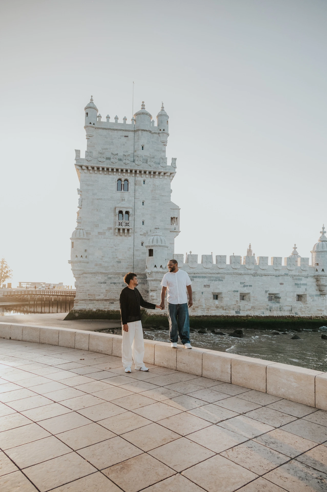 The couple holding hands while standing at different levels beside the low wall at Belém Tower during their couple photoshoot in Lisbon.