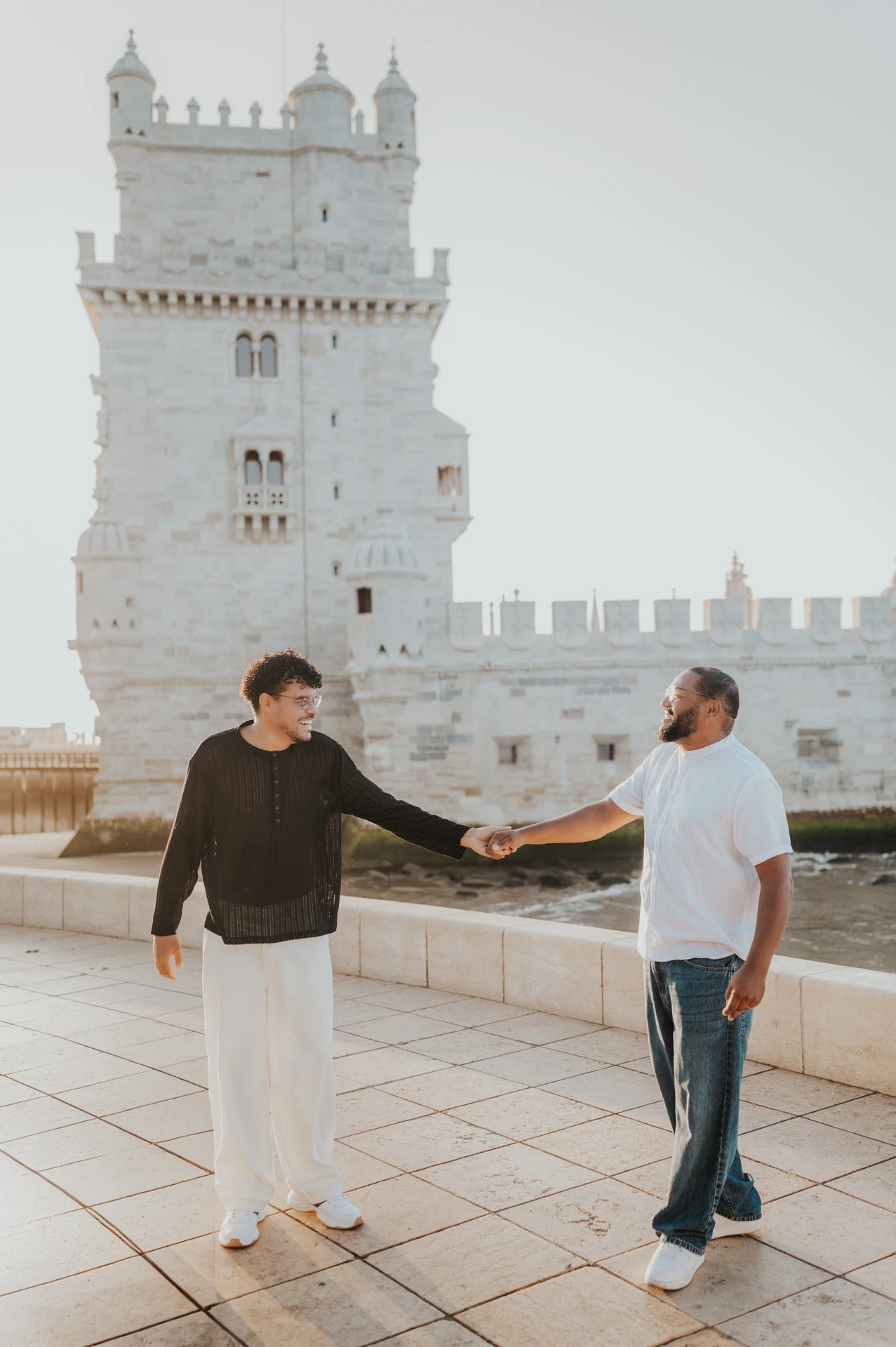 The couple standing apart with their joined hands extended between them beside Belém Tower during their couple photoshoot in Lisbon.