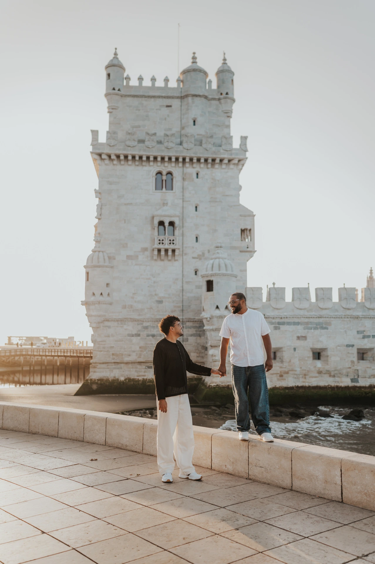 The couple holding hands with one partner on the low ledge and the other on the paving beside Belém Tower during their couple photoshoot in Lisbon.