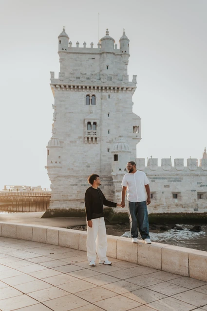 Holding hands at different levels | Lisbon couple photoshoot The couple holding hands with one partner on the low ledge and the other on the paving beside Belém Tower during their couple photoshoot in Lisbon.