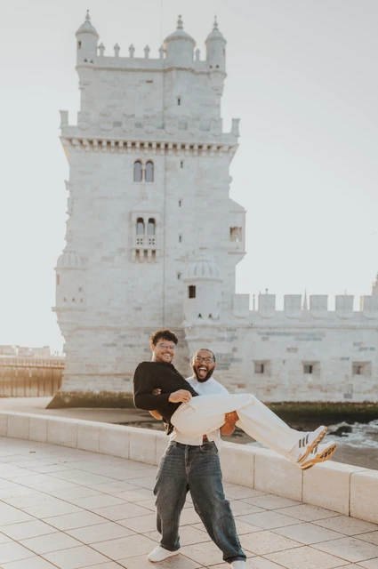 A playful lift beside the tower | Lisbon couple photoshoot One partner carrying the other horizontally in his arms beside the waterfront with Belém Tower behind them during their couple photoshoot in Lisbon.