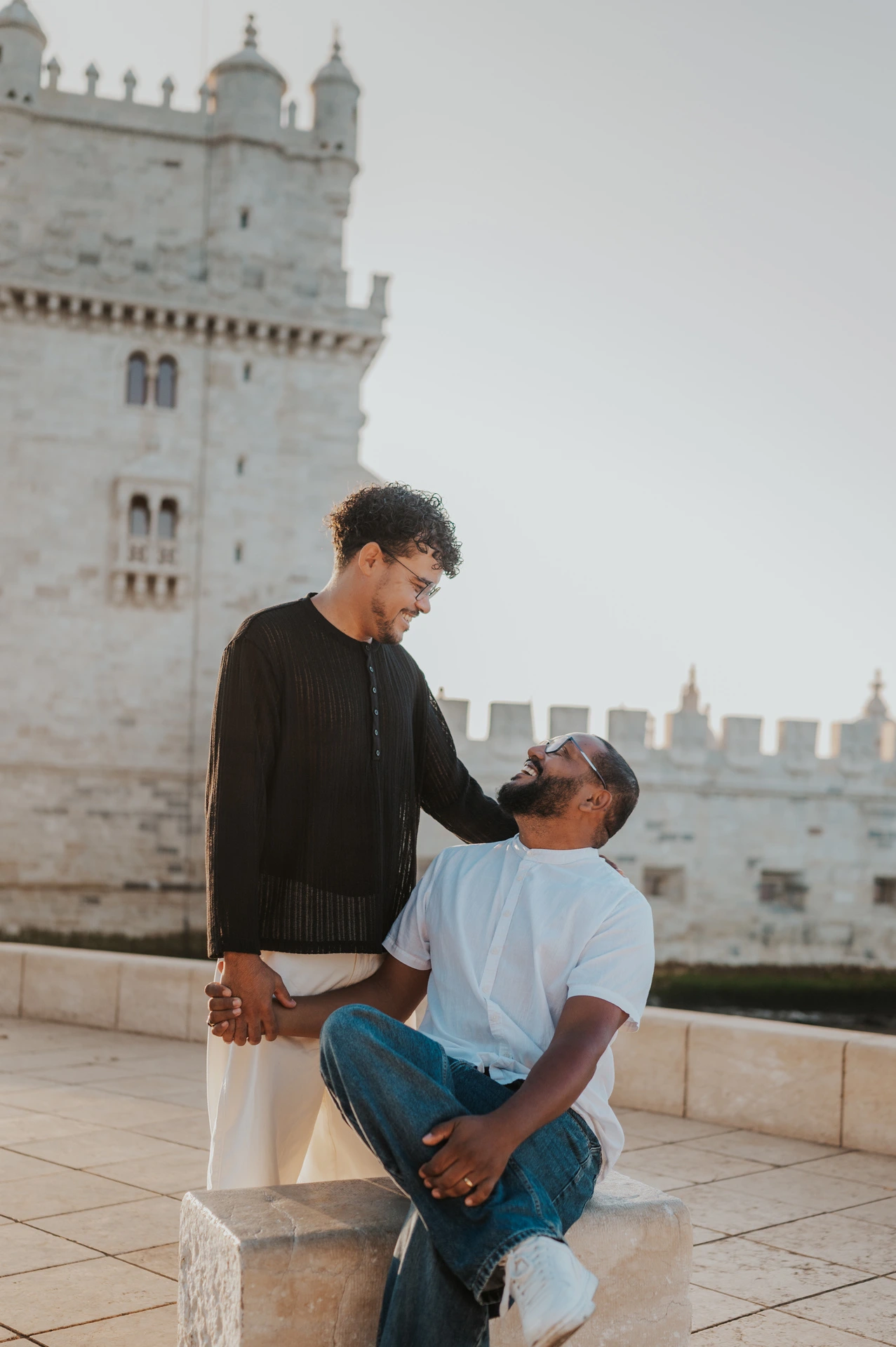 One partner seated on a stone block, looking up at the other with Belém Tower behind them during their couple photoshoot in Lisbon.