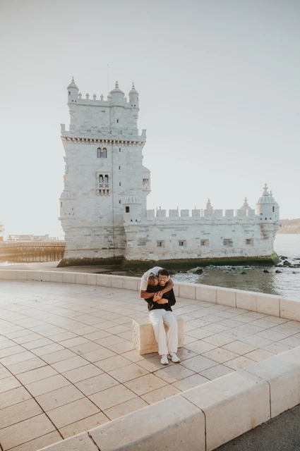 Leaning close on the waterfront ledge | Lisbon couple photoshoot The couple leaning close together beside a low waterfront ledge with Belém Tower behind them during their couple photoshoot in Lisbon.