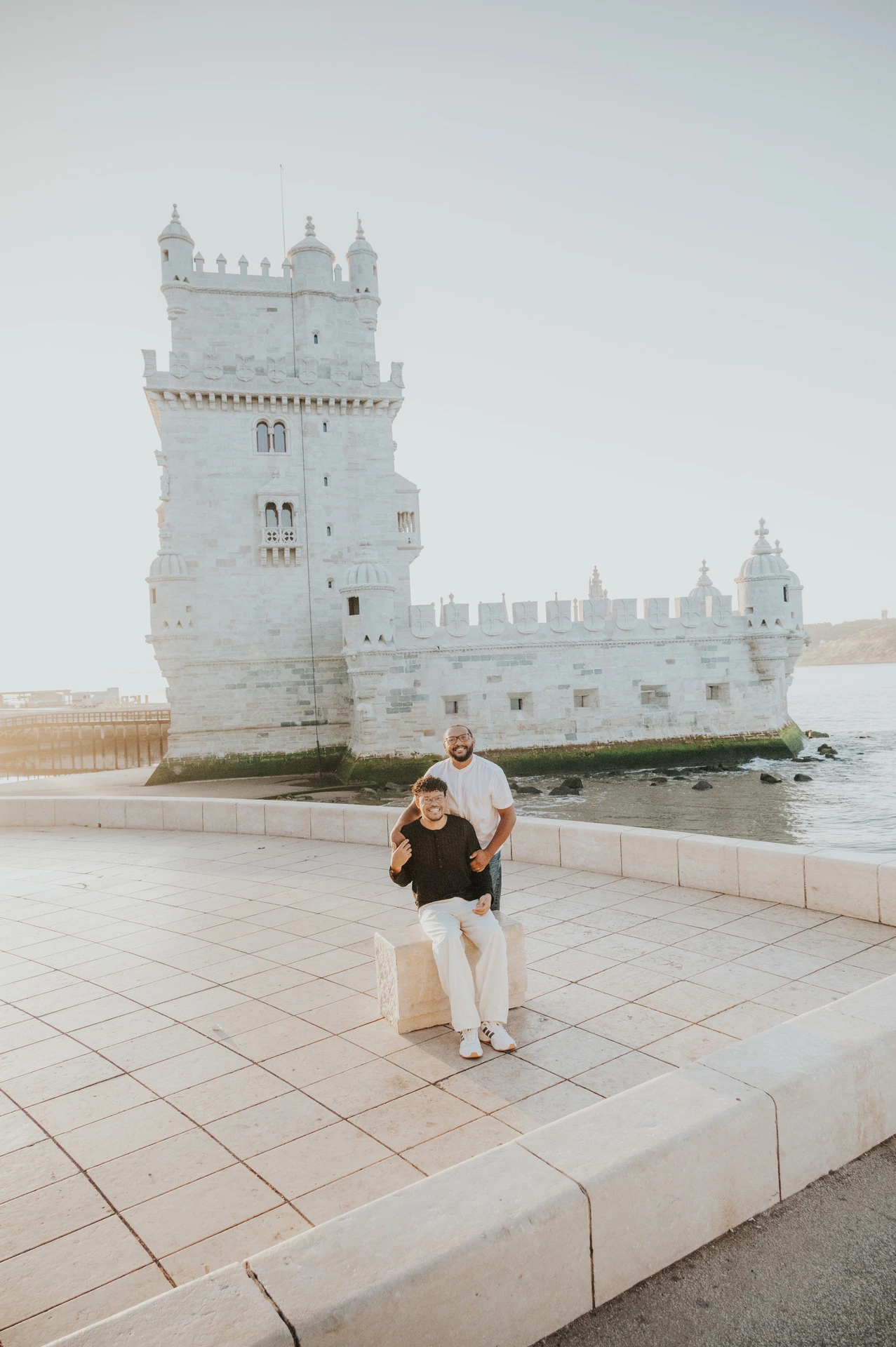 One partner sitting on the low ledge while the other stands behind him beside Belém Tower during their couple photoshoot in Lisbon.