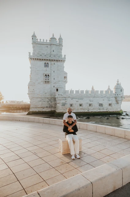 A seated portrait beside the water | Lisbon couple photoshoot One partner sitting on a low stone ledge with the other standing behind him and Belém Tower in the background during their couple photoshoot in Lisbon.