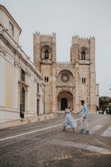 The couple holding hands on the cobbled street in front of a stone church with twin towers during their couple photoshoot in Lisbon.