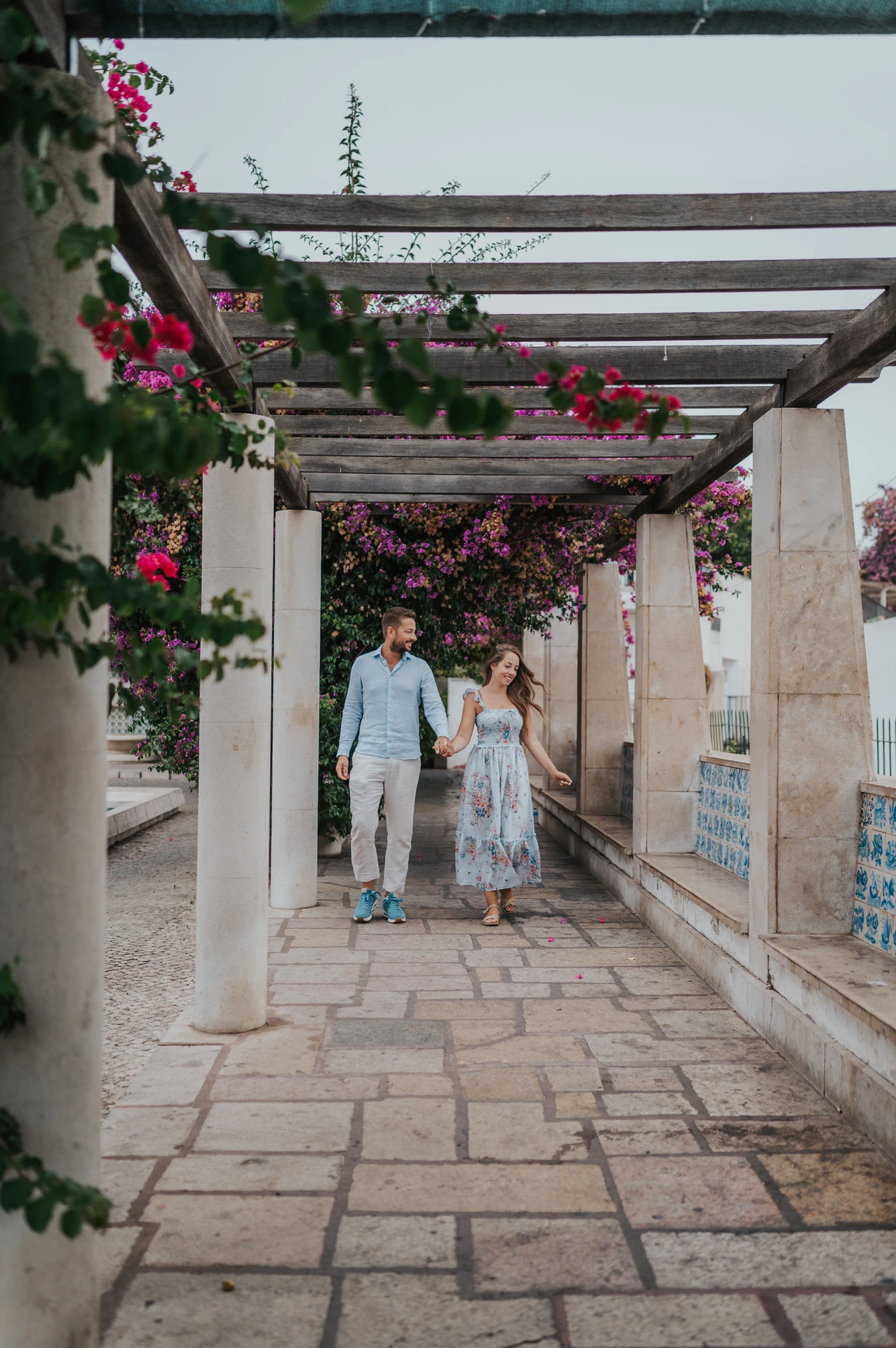 The couple walking hand in hand beneath the flower-covered wooden pergola at Santa Luzia during their couple photoshoot in Lisbon.