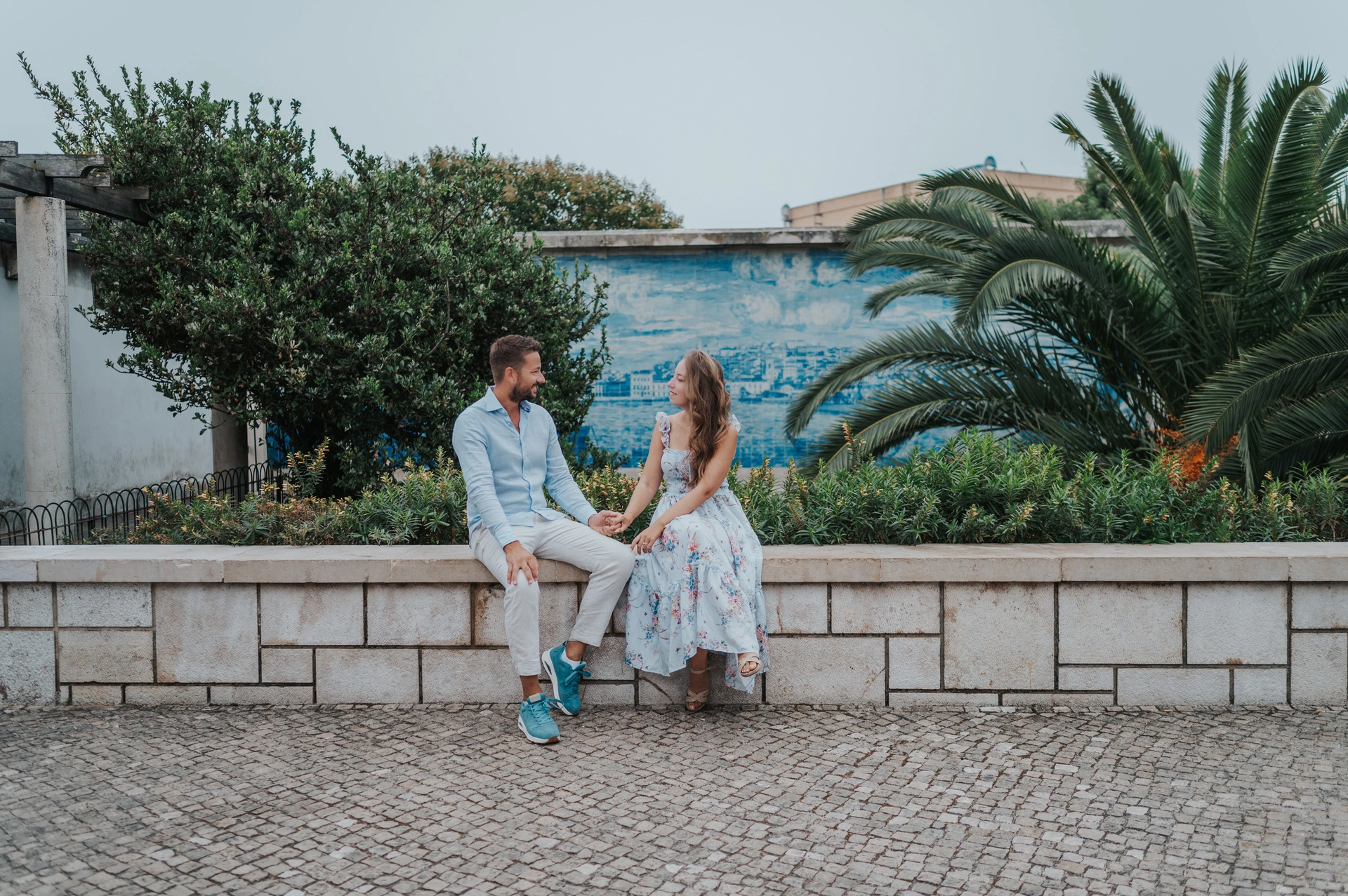 Together beside Santa Luzia’s blue tiles | Lisbon couple photoshoot The couple seated together on the garden wall with blue tiles and palms at Santa Luzia during their couple photoshoot in Lisbon.