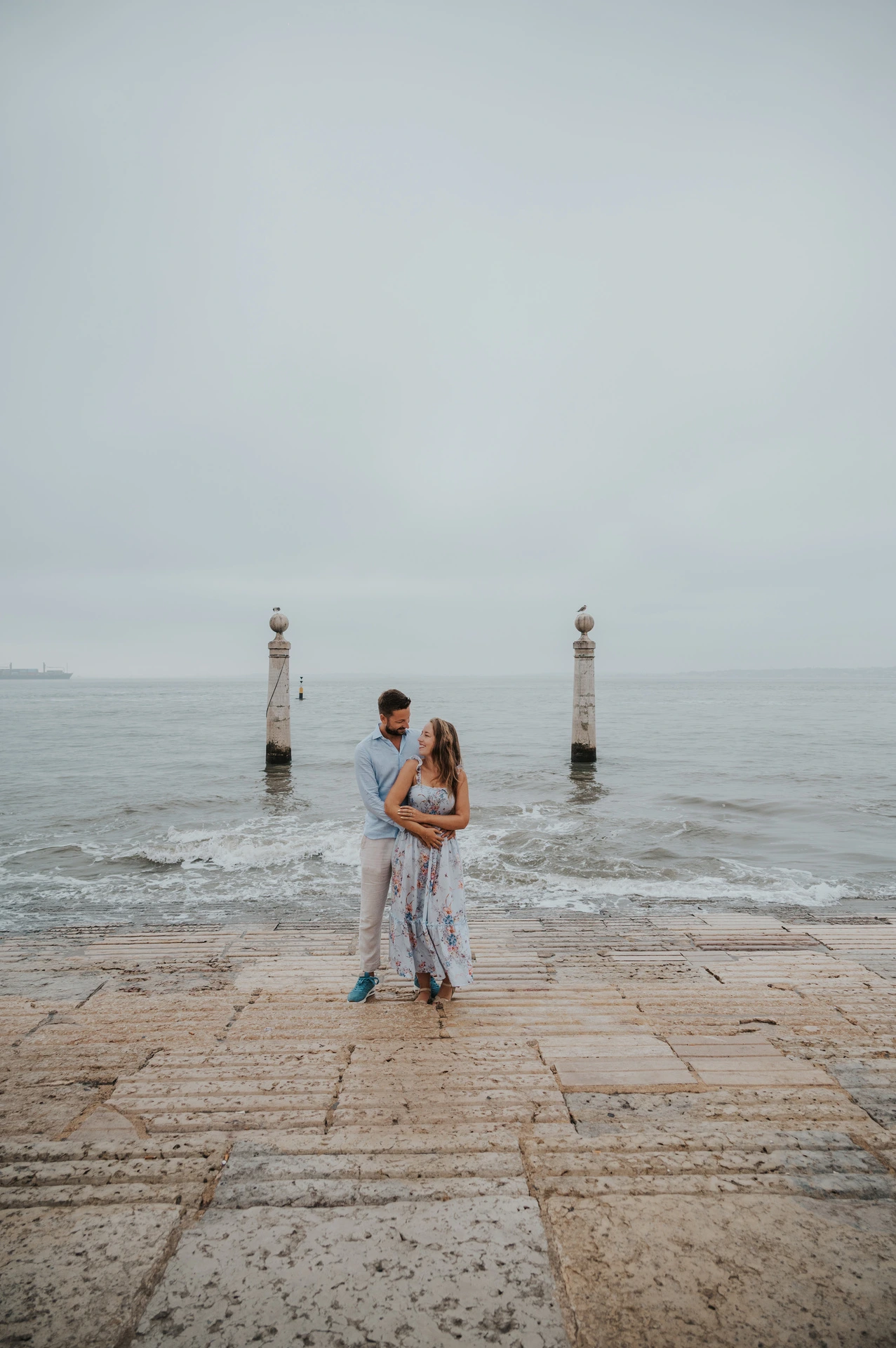 The couple leaning close together on the waterfront steps between two stone columns near Praça do Comércio during their couple photoshoot in Lisbon.