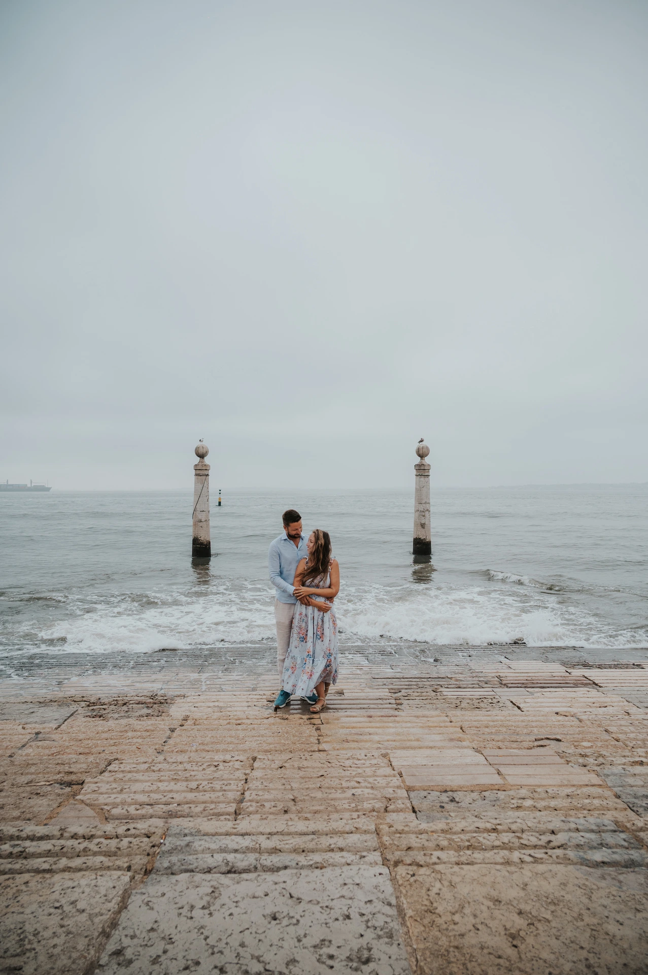 A full-length portrait of the couple embracing on the waterfront steps with waves behind them near Praça do Comércio during their couple photoshoot in Lisbon.