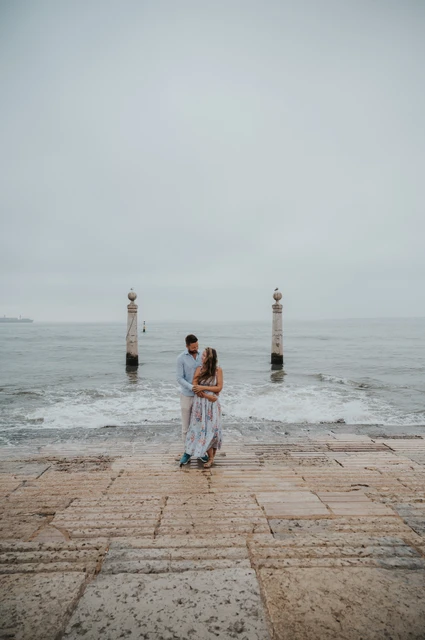 An embrace beside the waterfront waves | Lisbon couple photoshoot A full-length portrait of the couple embracing on the waterfront steps with waves behind them near Praça do Comércio during their couple photoshoot in Lisbon.