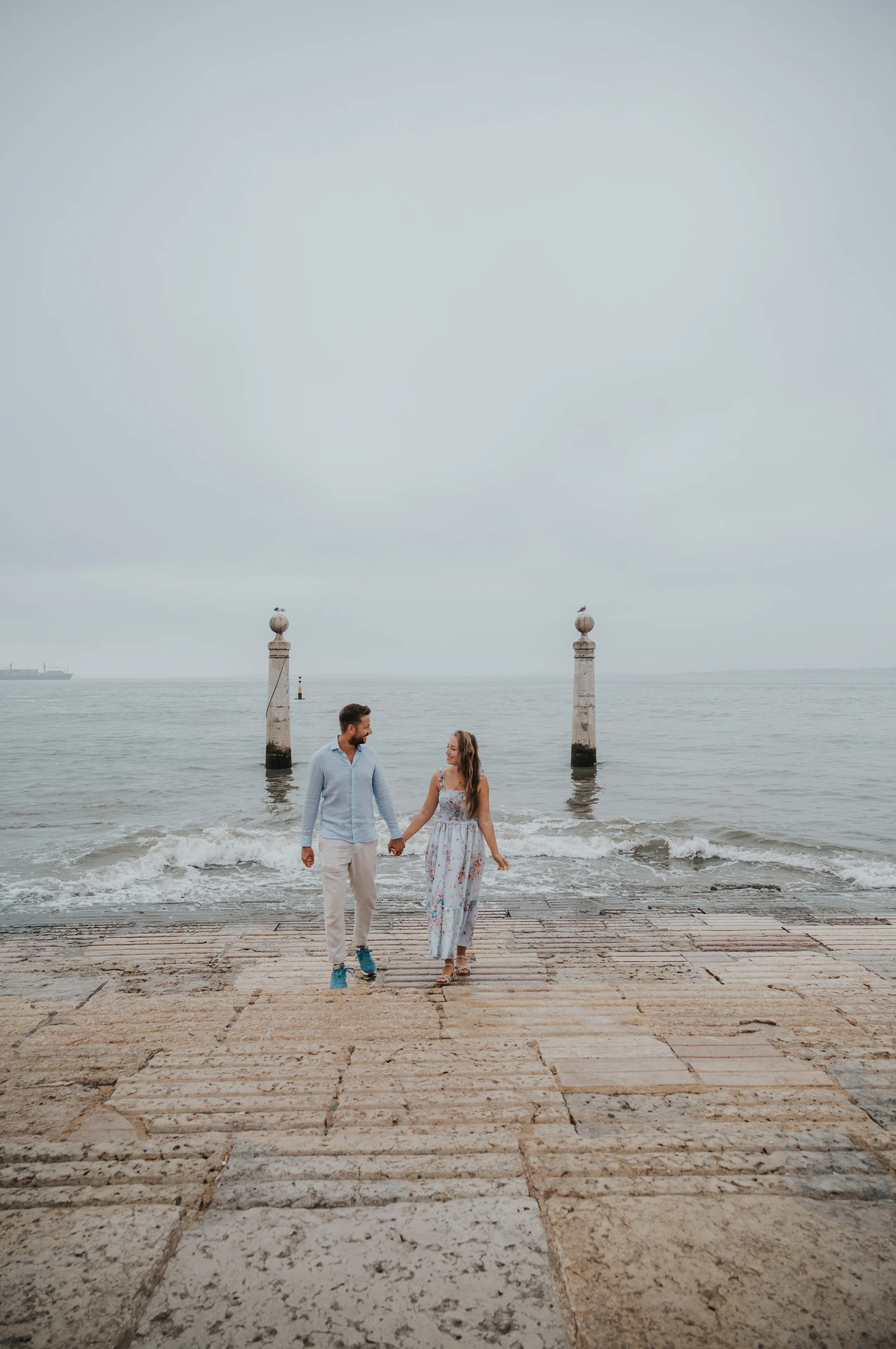 The couple standing apart with hands joined on the waterfront steps near Praça do Comércio during their couple photoshoot in Lisbon.