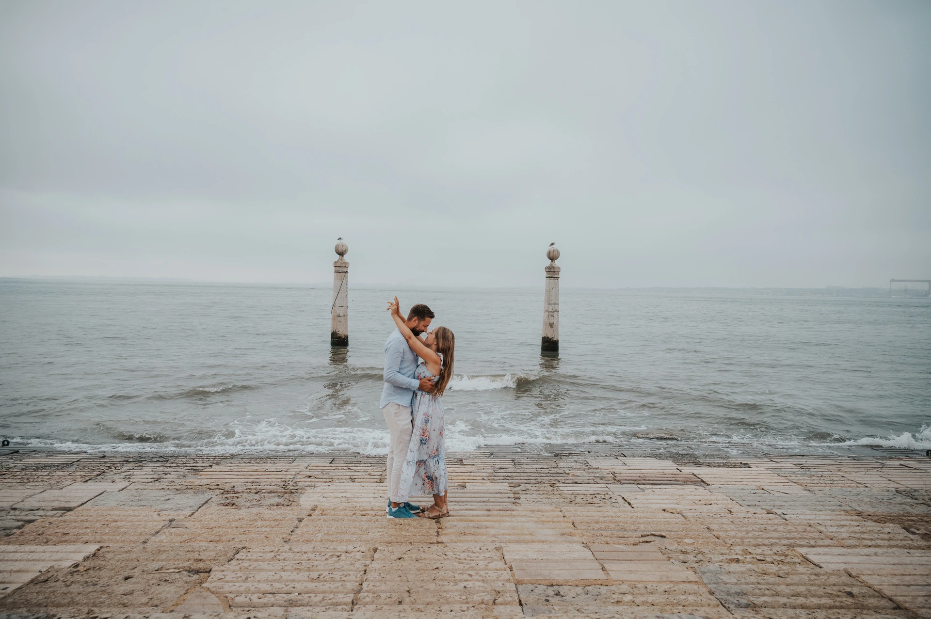 A wide portrait of the couple embracing with her arm raised beside the water near Praça do Comércio during their couple photoshoot in Lisbon.