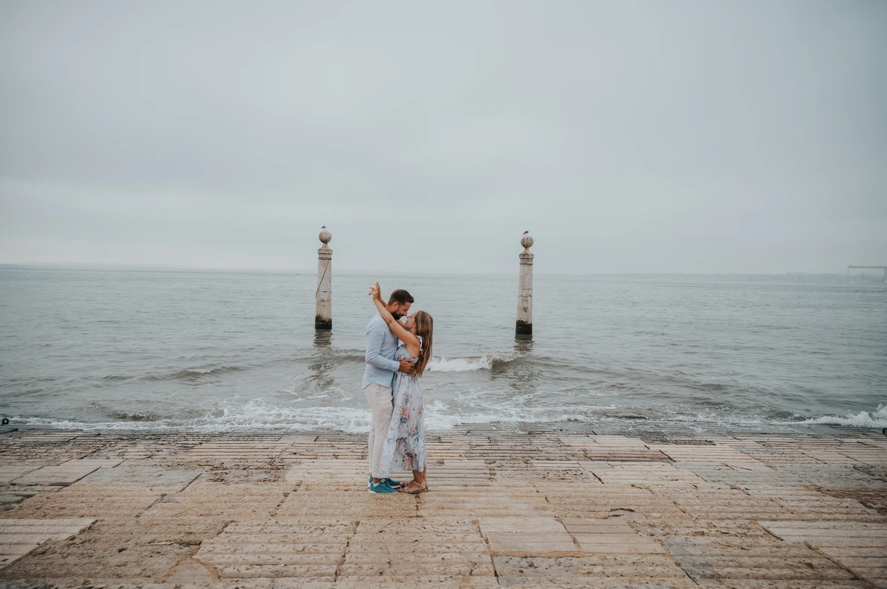 A wide embrace beside the river | Lisbon couple photoshoot A wide portrait of the couple embracing with her arm raised beside the water near Praça do Comércio during their couple photoshoot in Lisbon.
