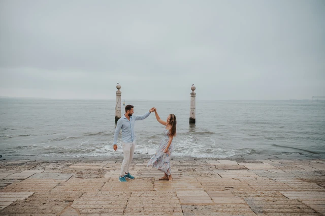 The couple raising their joined hands for a turn beside the waves near Praça do Comércio during their couple photoshoot in Lisbon.
