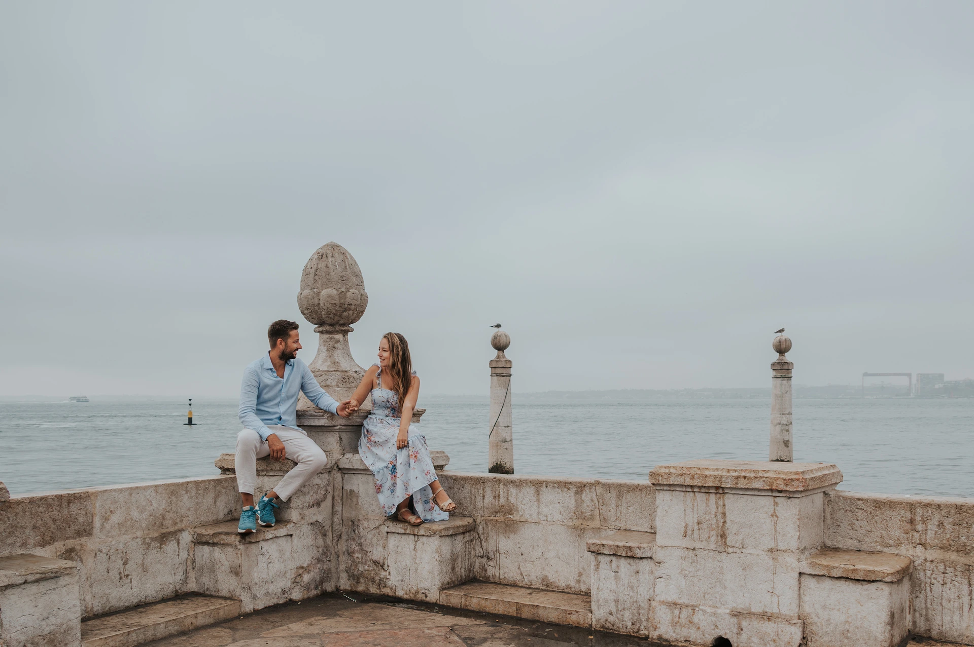 A shared glance above the water | Lisbon couple photoshoot The couple sitting on the stone waterfront wall and looking at each other with the river behind them during their couple photoshoot in Lisbon.
