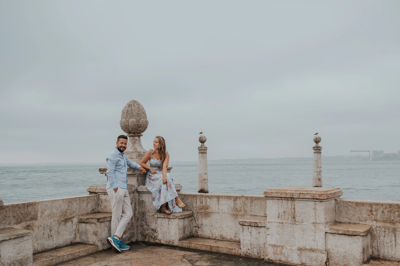 A portrait beside the stone waterfront wall | Lisbon couple photoshoot The woman seated on the stone waterfront wall with her partner standing beside her during their couple photoshoot in Lisbon.