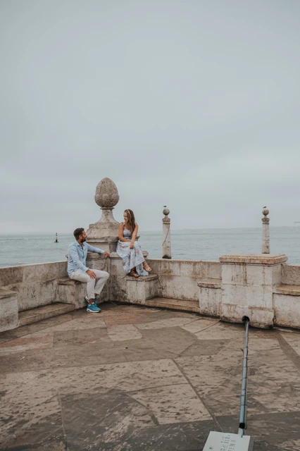 Seated together at the waterfront | Lisbon couple photoshoot A vertical portrait of the couple sitting together on the stone waterfront wall during their couple photoshoot in Lisbon.