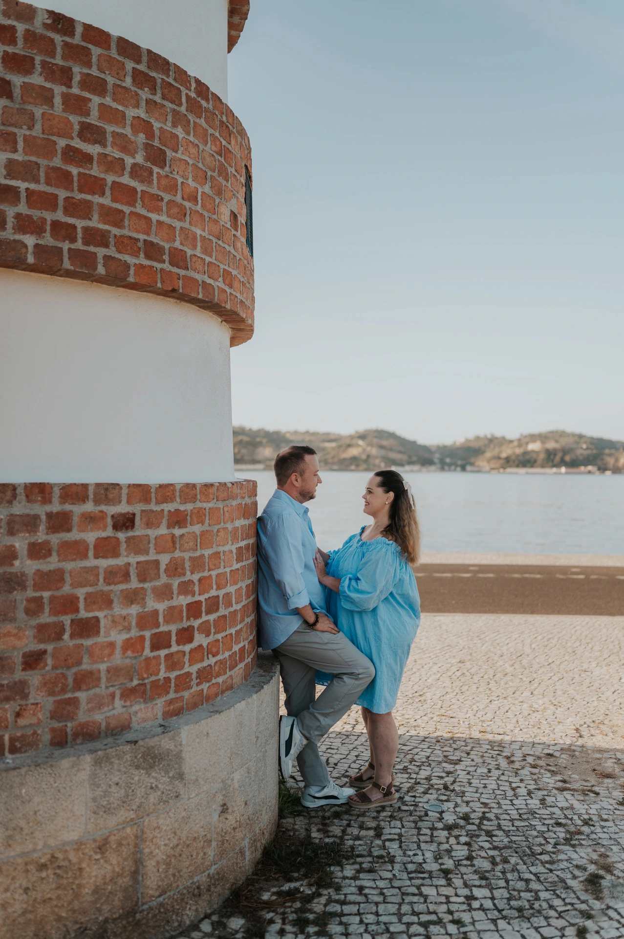 The couple leaning close together beside a curved red-brick and white wall on the waterfront during their engagement photoshoot in Lisbon.