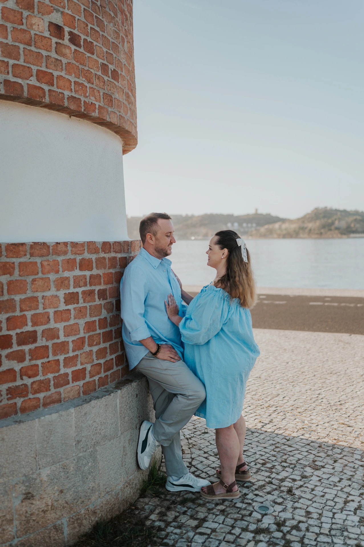 The couple leaning close together beside a curved red-brick and white wall on the waterfront during their engagement photoshoot in Lisbon.