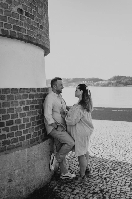 Together beside the curved waterfront wall in black and white | Lisbon engagement photoshoot Black-and-white photograph: the couple leaning close together beside a curved red-brick and white wall on the waterfront during their engagement photoshoot in Lisbon.