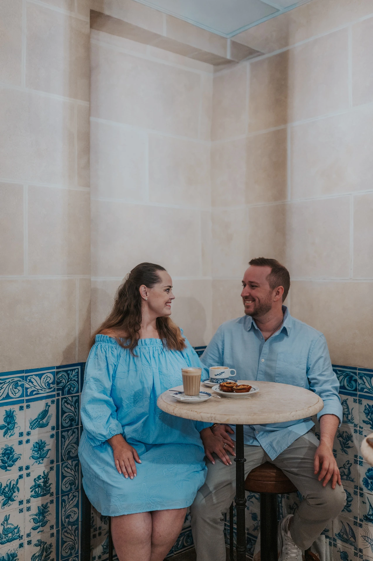 The couple smiling at one another over coffee and pastries at a small tiled café table during their engagement photoshoot in Lisbon.