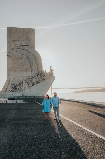The couple holding hands on the waterfront with the Monument to the Discoveries behind them during their engagement photoshoot in Lisbon.