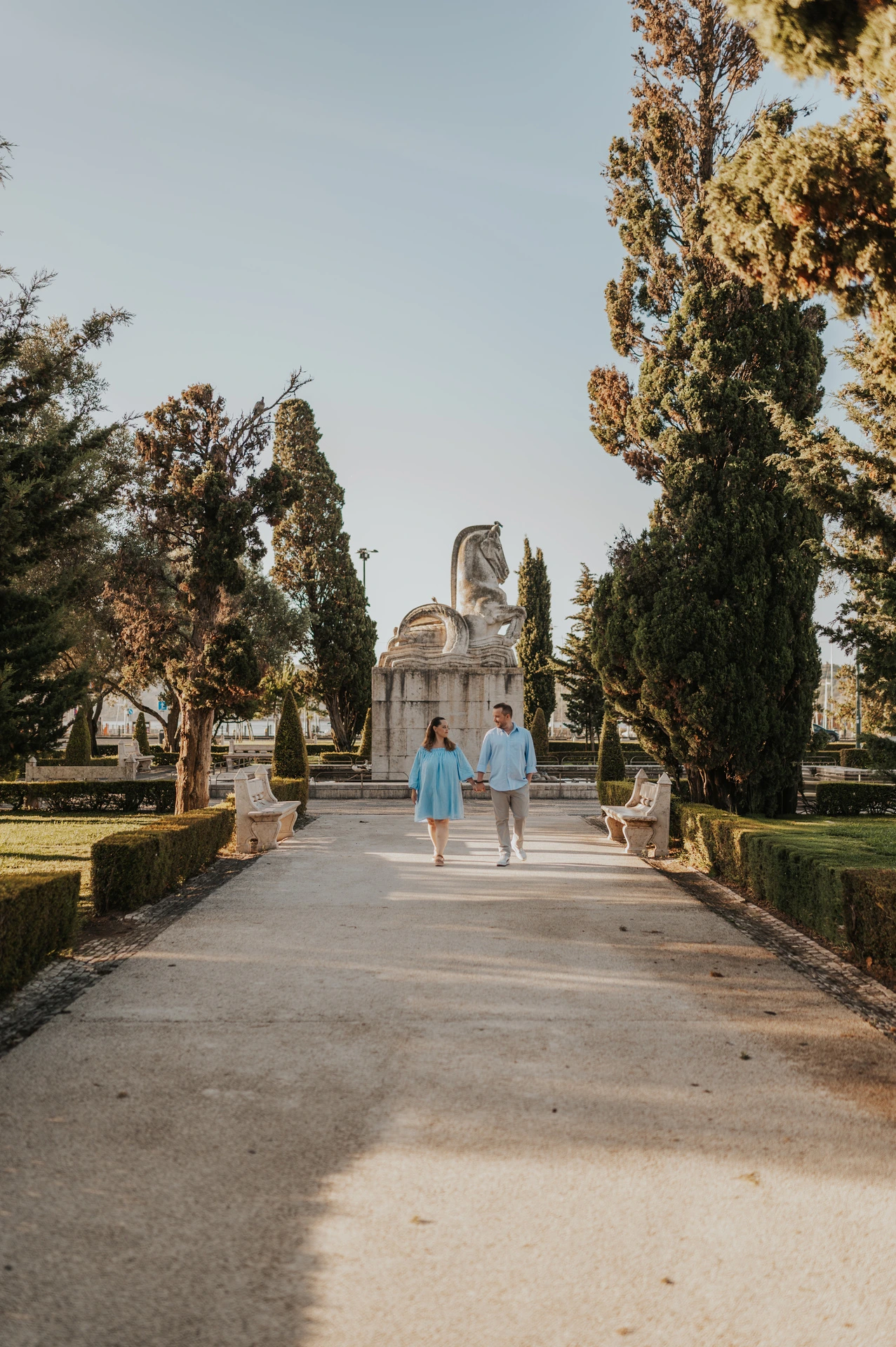 The couple walking hand in hand along the garden path between trees and hedges during their engagement photoshoot in Lisbon.