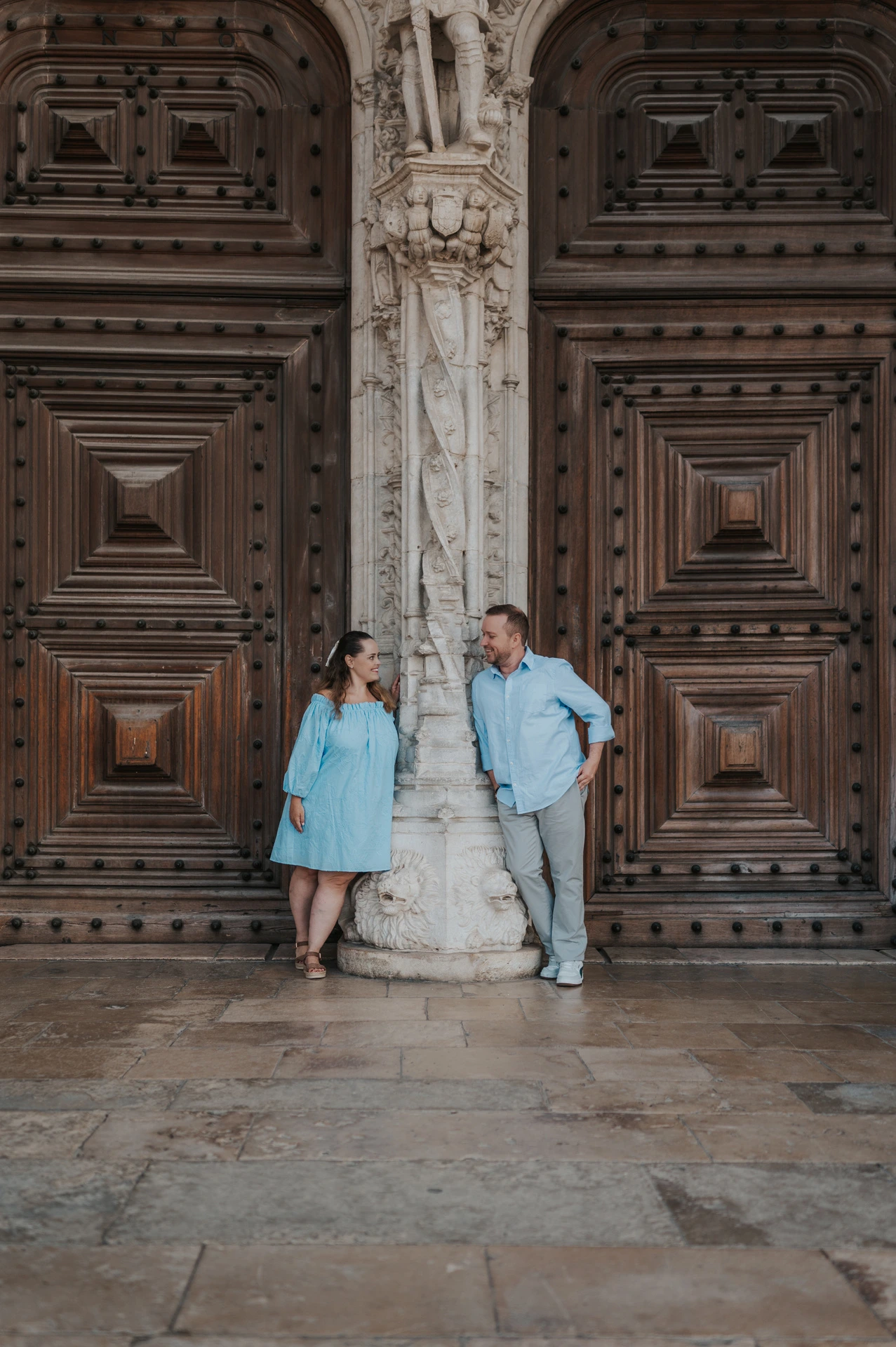 The couple leaning on either side of a carved stone column between the monastery’s wooden doors during their engagement photoshoot in Lisbon.