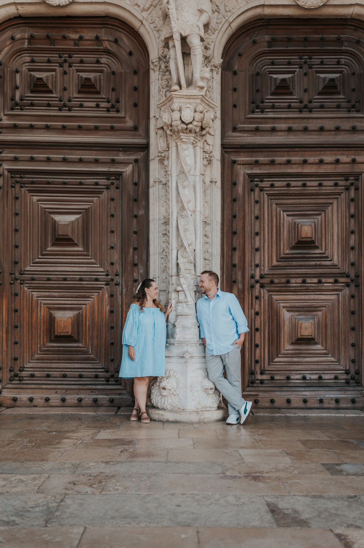 The couple leaning on either side of a carved stone column between the monastery’s wooden doors during their engagement photoshoot in Lisbon.