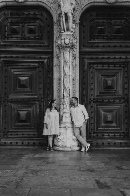 A portrait between the monastery doors in black and white | Lisbon engagement photoshoot Black-and-white photograph: the couple leaning on either side of a carved stone column between the monastery’s wooden doors during their engagement photoshoot in Lisbon.