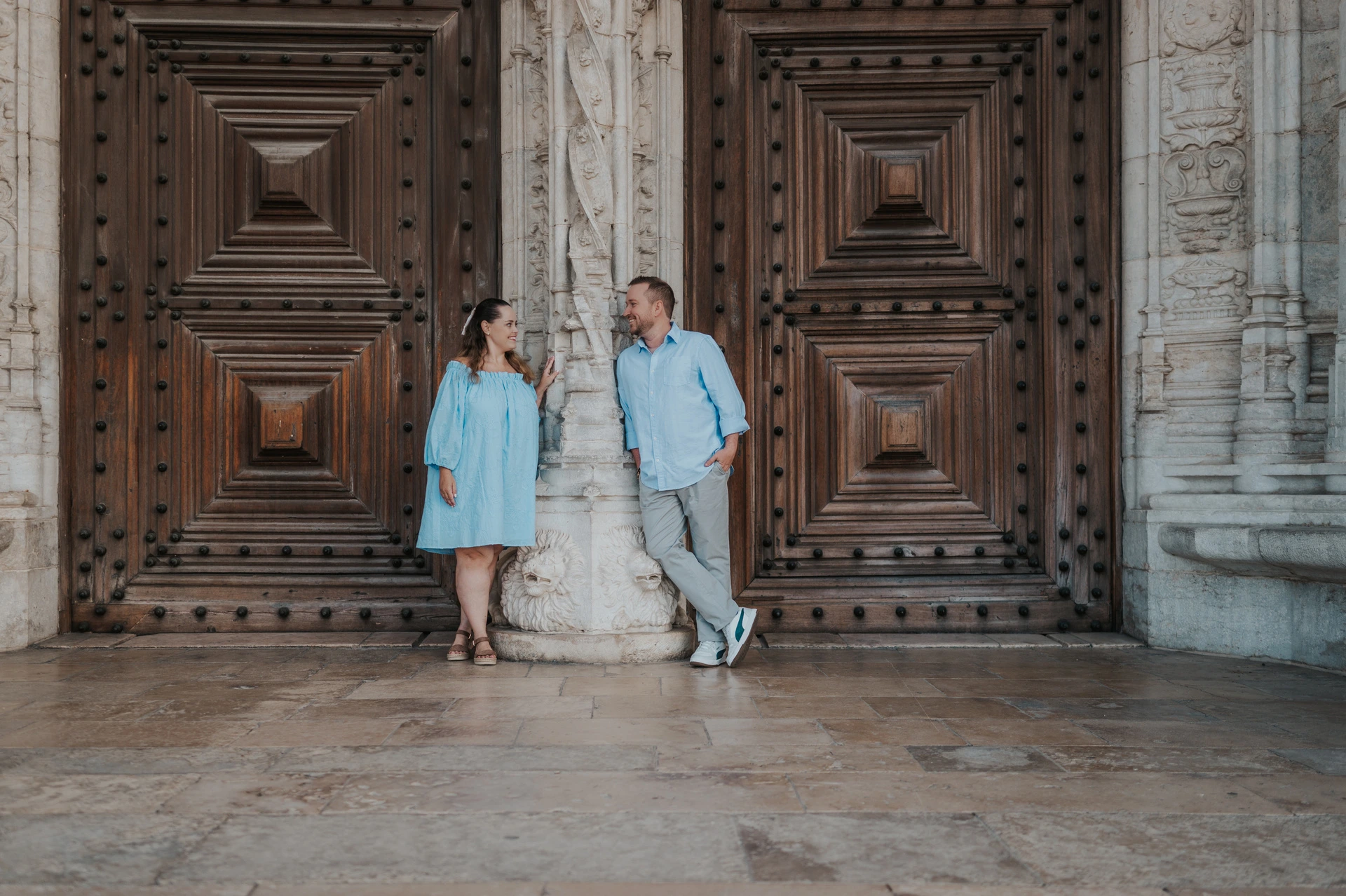 The couple leaning on either side of a carved stone column between the monastery’s wooden doors during their engagement photoshoot in Lisbon.