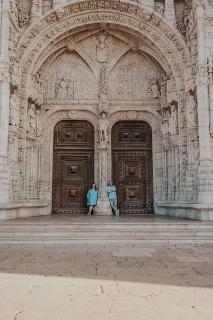 Beneath the monastery’s carved entrance | Lisbon engagement photoshoot A wide view of the couple beneath the ornate entrance of Jerónimos Monastery during their engagement photoshoot in Lisbon.