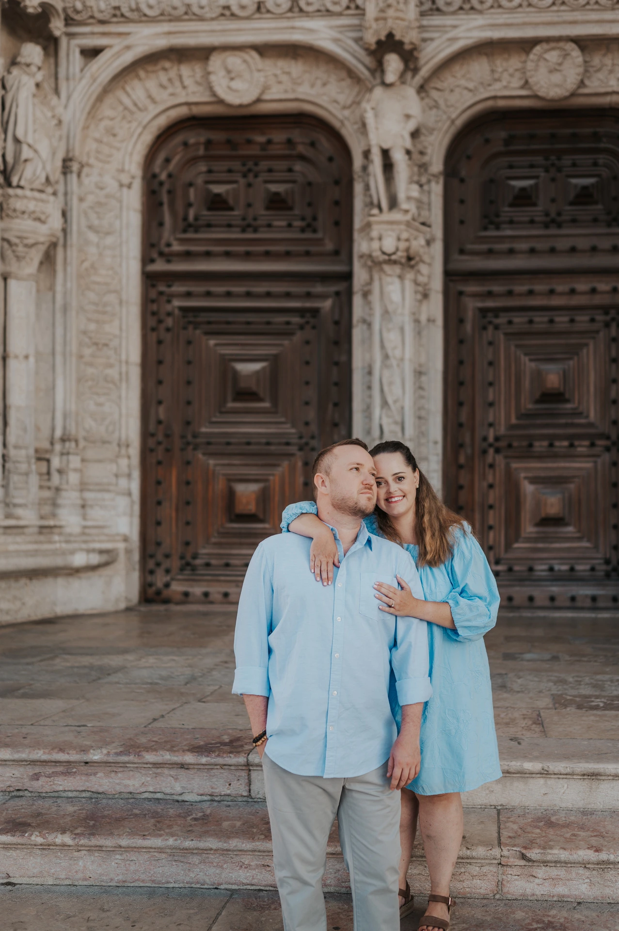 The woman with an arm around her partner’s shoulder as they smile outside the monastery during their engagement photoshoot in Lisbon.