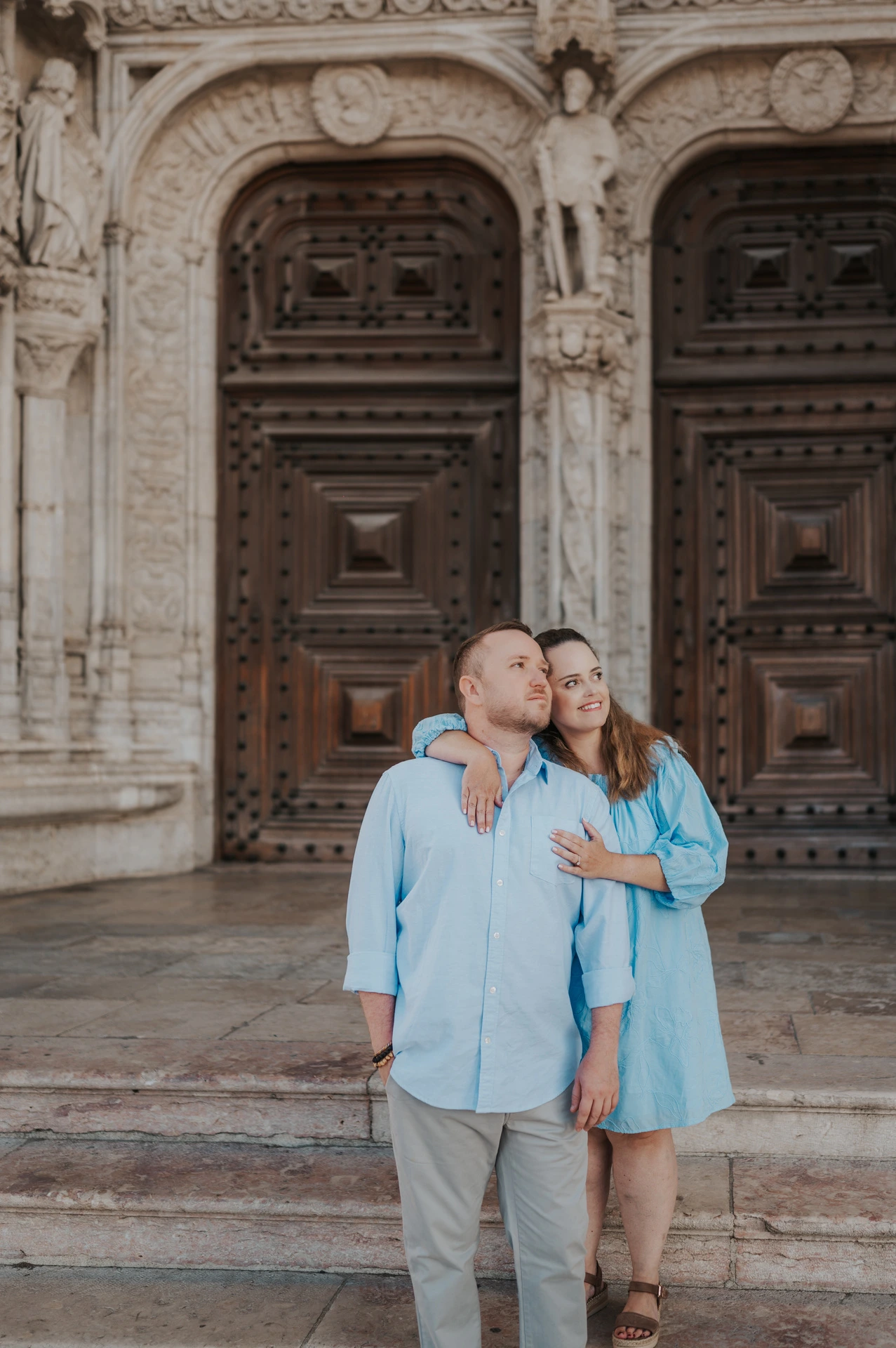The woman with an arm around her partner’s shoulder as they smile outside the monastery during their engagement photoshoot in Lisbon.