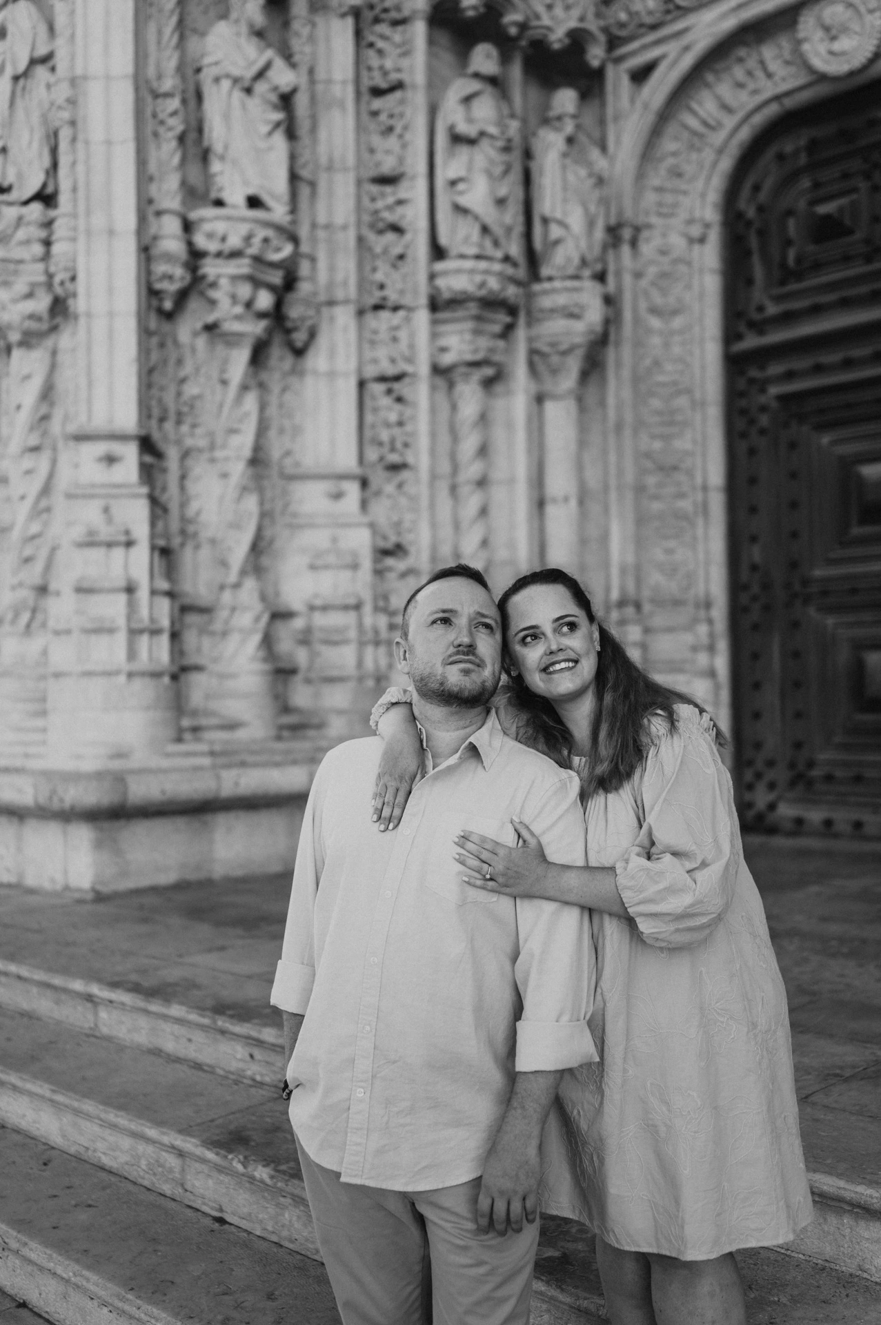 Black-and-white photograph: the woman with an arm around her partner’s shoulder as they smile outside the monastery during their engagement photoshoot in Lisbon.