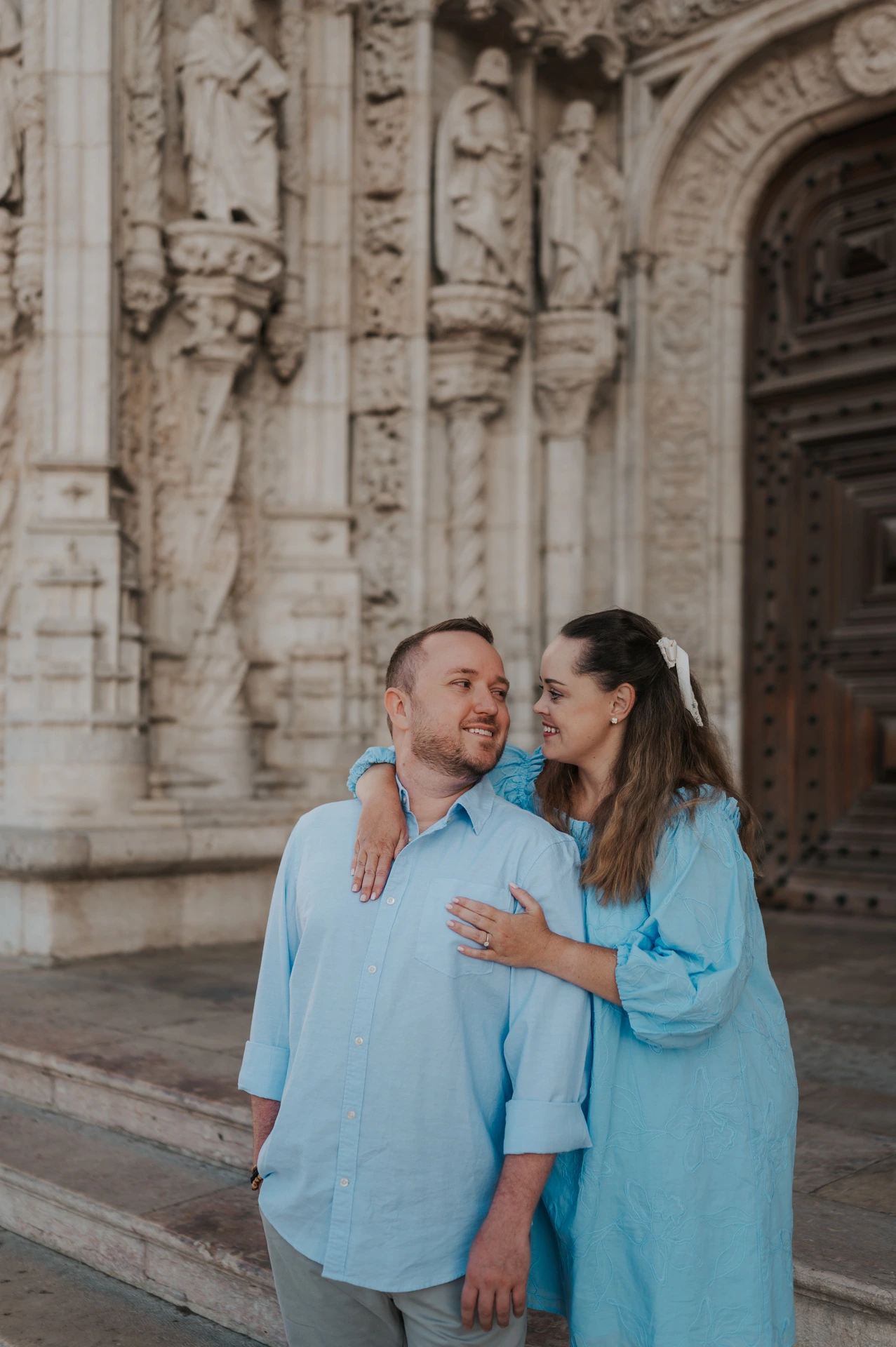 The woman with an arm around her partner’s shoulder as they smile outside the monastery during their engagement photoshoot in Lisbon.