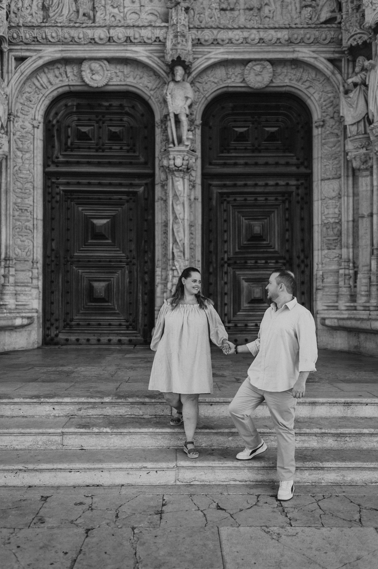 Black-and-white photograph: the couple holding hands and moving together on the steps outside Jerónimos Monastery during their engagement photoshoot in Lisbon.