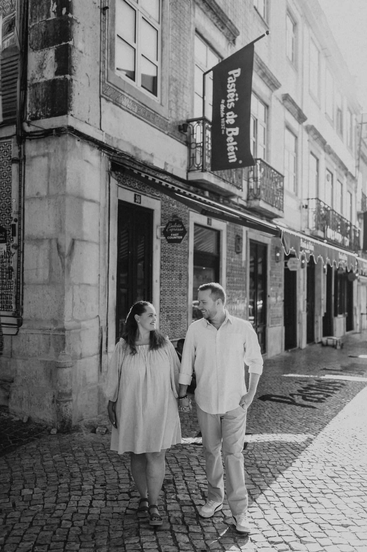 Black-and-white photograph: the couple walking hand in hand outside Pastéis de Belém during their engagement photoshoot in Lisbon.