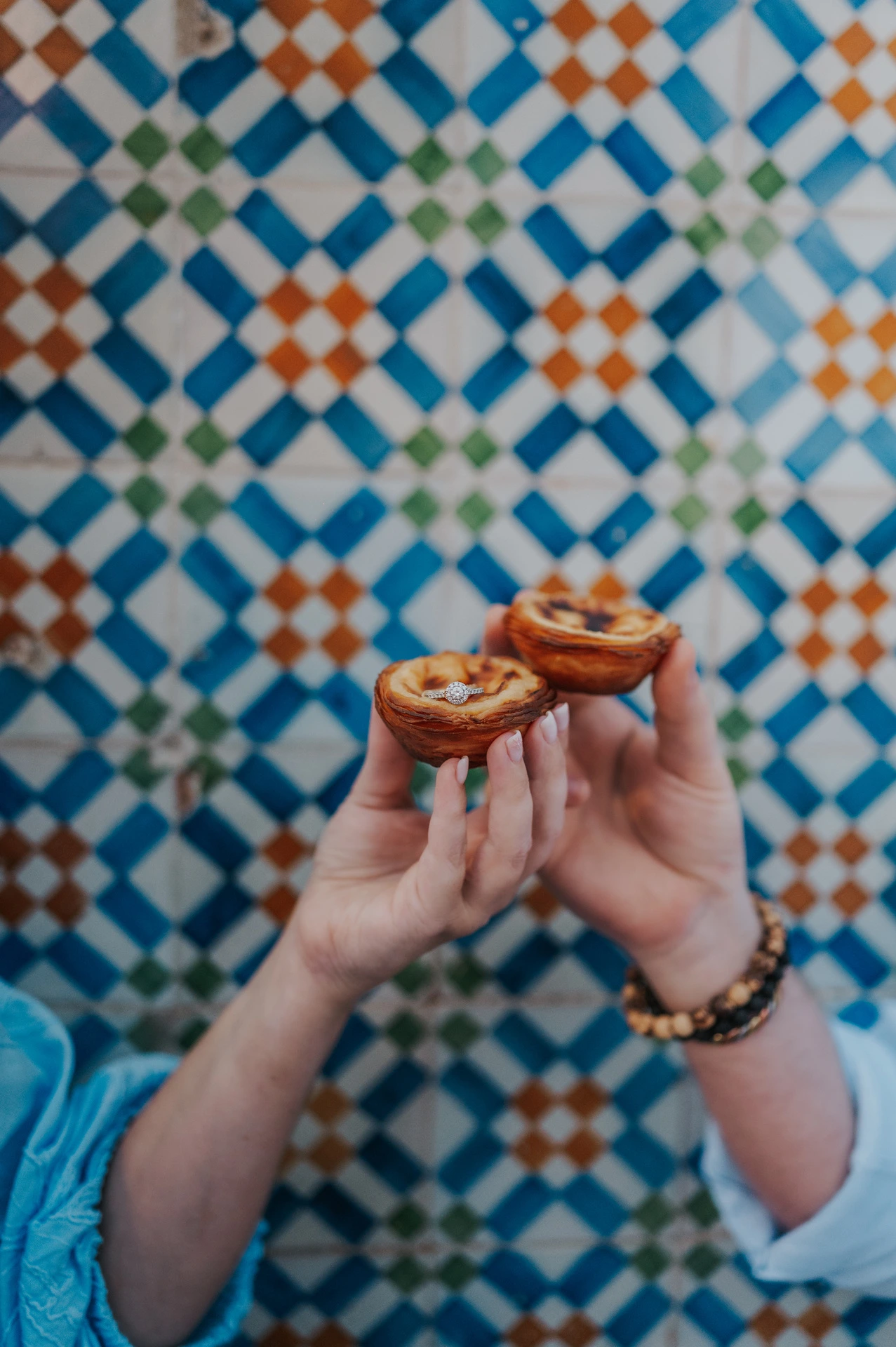 The couple holding two custard tarts together in front of the colourful patterned tiles during their engagement photoshoot in Lisbon.