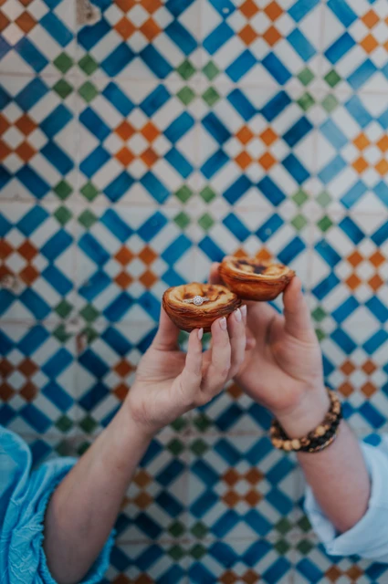 Two pastéis held up against the tiles | Lisbon engagement photoshoot The couple holding two custard tarts together in front of the colourful patterned tiles during their engagement photoshoot in Lisbon.