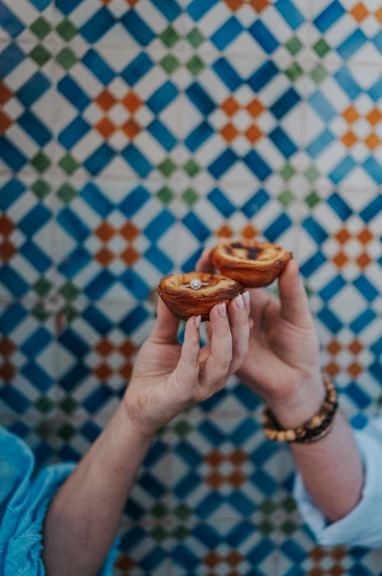 Two pastéis held up against the tiles | Lisbon engagement photoshoot The couple holding two custard tarts together in front of the colourful patterned tiles during their engagement photoshoot in Lisbon.
