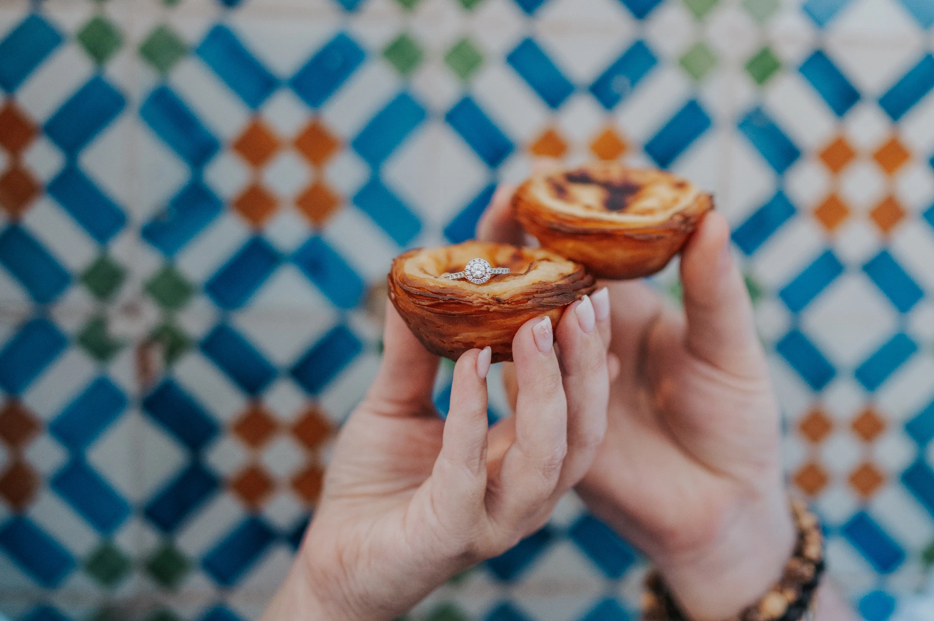 The couple holding two custard tarts together in front of the colourful patterned tiles during their engagement photoshoot in Lisbon.