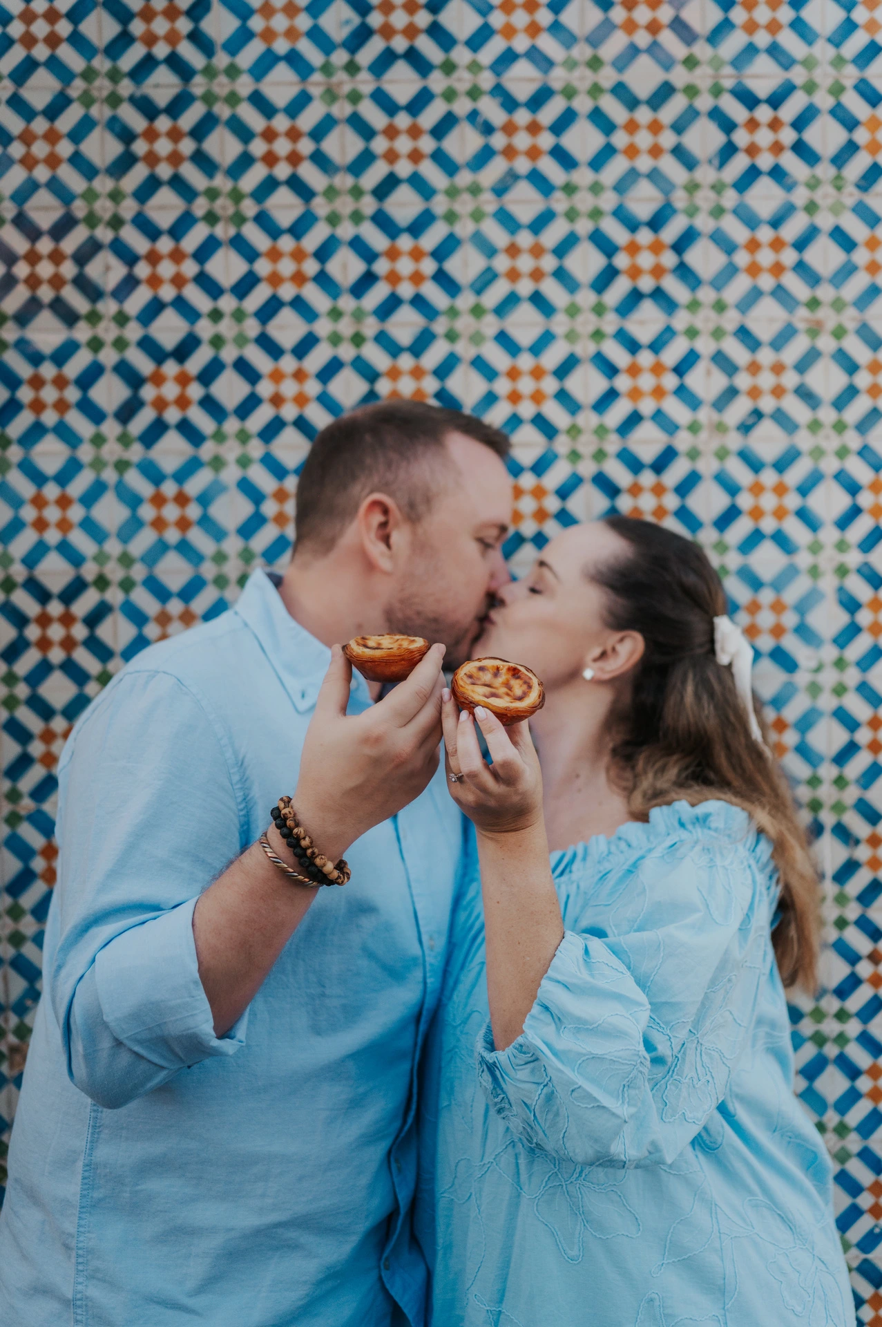 The couple leaning together with custard tarts held close to their faces against the patterned tile wall during their engagement photoshoot in Lisbon.